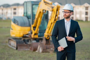 Takım elbiseli ve kasklı bir Buider. İnşaat yatırımcısı. İnşaat alanının önündeki yatırımcı. Başarılı bir yatırımcı. İnşaatta takım elbiseli yakışıklı ve kasklı bir adam.