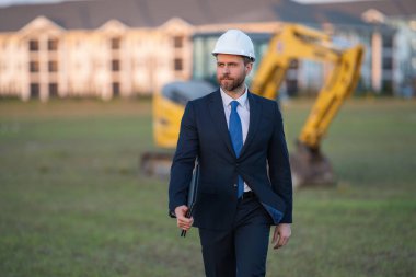 İnşaat alanında kask ve takım elbiseli bir inşaat yöneticisi. İnşaat müdürü ya da yönetici evin önünde şapka takıyor. Kazıcının yanındaki inşaat müdürü. Yenileme