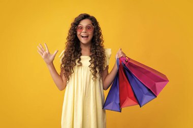Sarı arka planda alışveriş torbaları olan güzel moda kızı. Alışveriş ve moda alışverişi. Alışveriş çantalı çocuk. Şaşırmış yüz, şaşırmış duygular.