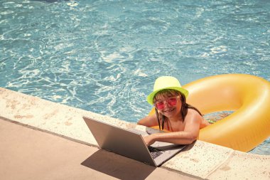Çocuk yüzme havuzunda dizüstü bilgisayarla çalışıyor. Yaz çevrimiçi teknoloji. Yaz tatilinde tropikal suda dinlenen bir gezgin. Seyahat, dizüstü bilgisayar kullanan çocuk.
