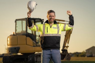 Man builder near excavator. Builder worker with excavator. Builder in helmet. Worker in hardhat. Portrait mechanical worker in construction helmet. Engineer builder foreman or repairman