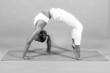 Kız esniyor, köprü pozu veriyor. Sportif kız, yoga minderi üzerinde egzersiz yaparken, bacaklarını yukarı kaldırıyor, arka planda gri bir alan var.