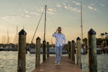 Yatın yanında zengin bir iş adamı var. Moda erkek modelinin açık havada portresi. Moda erkeği yatların yanında poz veriyor. Kentsel tarzda bir adam. Yat kulübündeki zengin adam. Deniz manzaralı ve lüks gemileri olan körfez adamı.