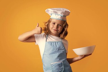Funny kid chef cook with kitchen plate, studio portrait. Child chef cook prepares food on isolated studio background. Kids cooking. Teen boy with apron and chef hat preparing a healthy meal