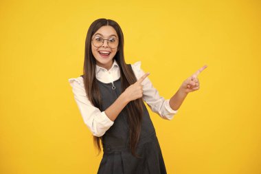 Look at advert. Teenager child points aside shows blank copy space for text promo idea presentation, poses against yellow background. Excited face, cheerful emotions of teenager girl