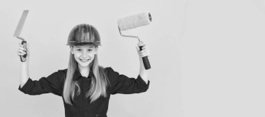 glad teen child in builder hard hat with paint roller and spatula on yellow wall. Child builder in helmet horizontal poster design. Banner header, copy space