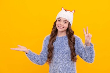 Kalın giysiler giyen çocuk portresi. Olumlu duygularını ifade et. Kış modası. Gülümseyen çocuk fotokopi alanında ürün sunuyor. Kadın manken. Sarı arka planda örgü elbiseli genç kız..