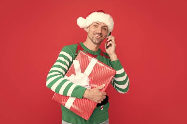 Neşeli elf telefonda konuşuyor. Noel Baba şapkalı adam telefonda kırmızı arka planda mevcut kutusuyla selamlaşıyor. yeni yıl.