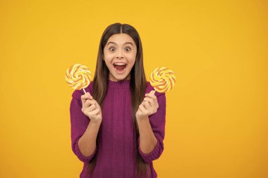 Excited face. Child eat lollipop popsicle over yellow isolated background. Yummy caramel, candy shop. Amazed expression, cheerful and glad
