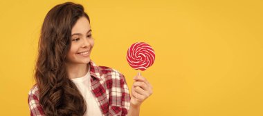 Uzun kıvırcık saçlı, kareli tişörtlü neşeli çocuk sarı arka planda lolipop karamel şeker, diş sağlığı.