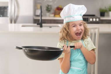 Çocuk aşçı mutfakta tavayla aşçılık yapıyor. Aşçı çocuk mutfakta yemek yapıyor. Çocuk aşçı mutfakta yemek hazırlar. Çocuklar yemek yapıyor. Önlüğü ve aşçı şapkası olan genç çocuk sağlıklı bir yemek hazırlıyor.