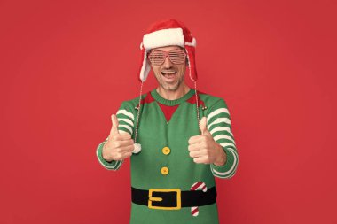 thumb up. surprised man in elf costume and party glasses. xmas guy in santa claus hat on red background. happy new year. merry christmas. clipart