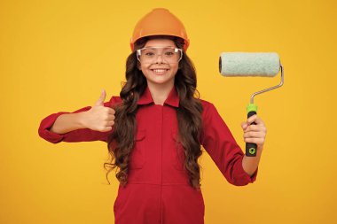 Kids renovation and building concept. Little girl in protective helmet and paint brush for painting walls isolated on yellow background. Happy face, positive and smiling emotions of teenager girl