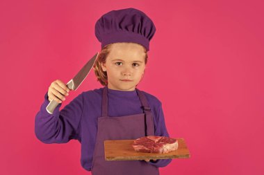 Child cook hold cutting board with meat beef steak and knife. Portrait of little child in uniform of cook. Chef boy isolated on studio background. Cute child to be a chef. Child dressed as a chef hat