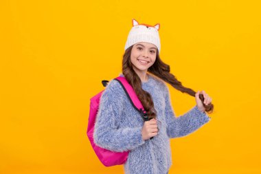 Sarı arka planda, şapkalı, uzun saçlı mutlu çocuk okul çantasını tutuyor..