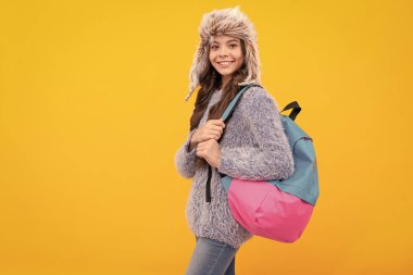 happy child in sweater and earflap hat with backpack on yellow background with copy space, school.