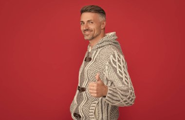 Kırmızı arka planda süveter giyen neşeli, kır saçlı adam baş parmağıyla jest yapıyor, erkek güzeli..