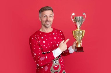 amazed xmas guy celebrating success on red background. happy new year. merry christmas. man in winter sweater point finger on champion cup. clipart