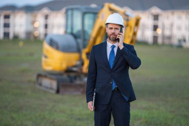 İnşaat işyeri sahibi. İnşaat alanında takım elbiseli ve şapkalı bir adam. İnşaat mühendisi kazıcının yanındaki evin önünde. Mimar, yönetici, yatırımcı, inşaat yöneticisi