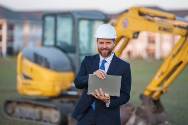İnşaat alanında kask ve takım elbiseli bir inşaat yöneticisi. İnşaat müdürü ya da yönetici evin önünde şapka takıyor. Kazıcının yanındaki inşaat müdürü. Yenileme