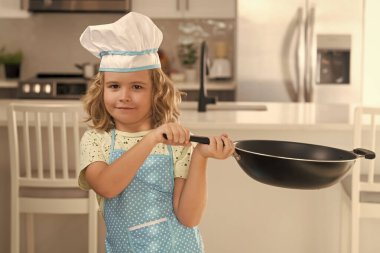 Çocuk aşçı mutfakta tavayla aşçılık yapıyor. Eğlenceli çocuklar mutfağı. Üniformalı aşçı şapkası ve önlük takan komik aşçı çocuk mutfakta yemek pişirdi. Çocuklar yemek hazırlıyor, kurabiye pişiriyor.