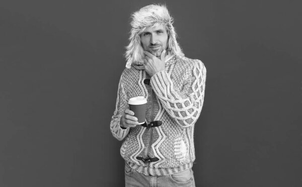 thinking hoary man drinking coffee. male fashion model hold cup. winter fashion. morning coffee in take away cup. grizzled guy in sweater and hat on red background.