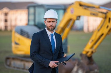 İnşaat işyeri sahibi. İnşaat alanında takım elbiseli ve şapkalı bir adam. İnşaat mühendisi kazıcının yanındaki evin önünde. Mimar, yönetici, yatırımcı, inşaat yöneticisi
