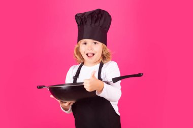 Aşçı çocuk tavayla aşçılık yapıyor. Çocuk fırın üniforması giyiyor ve aşçı şapkası takıyor. Mutfakta yemek hazırlıyor, stüdyo fotoğrafı çekiyor. Aşçılık, aşçılık ve çocuk maması konsepti.