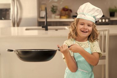 Çocuk aşçı mutfakta tavayla aşçılık yapıyor. Aşçı çocuk mutfakta aşçılık yapıyor. Mutfakta yemek pişiren sevimli çocuk. Sağlıklı malzemeler. Aşçı üniformalı küçük bir çocuğun portresi.