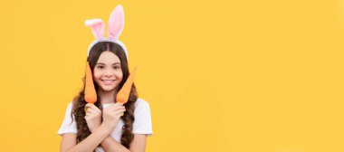 Tavşan kulaklı gülümseyen çocuk sarı arka planda havuç tutuyor. Paskalya çocuğu yatay posteri. Tavşan çocuğun web afişi, kopyalama alanı
