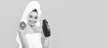 smiling kid in towel with grapefruit shampoo bottle on yellow background. Cosmetics and skin care for teenager child, poster design. Beauty kid girl banner with copy space