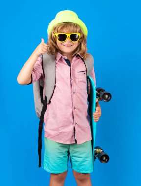 Mavi arka planda uzun tahta tutan çocuk. Pennyboard 'u olan çocuk. Moda şapkalı ve güneş gözlüklü neşeli küçük çocuğun stüdyo fotoğrafı.