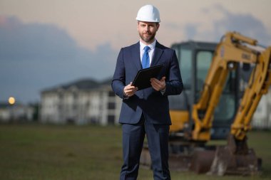 İnşaat alanında kask ve takım elbiseli bir inşaat yöneticisi. İnşaat müdürü ya da yönetici evin önünde şapka takıyor. Kazıcının yanındaki inşaat müdürü. Yenileme