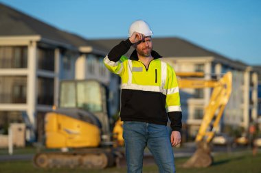 İnşaat alanında inşaat için kazı yapan bir inşaat işçisi. Binaların arka planında inşaat işçisi olan bir makine traktörü. Kazı işçisi. İnşaat alanında miğferi olan bir inşaatçı