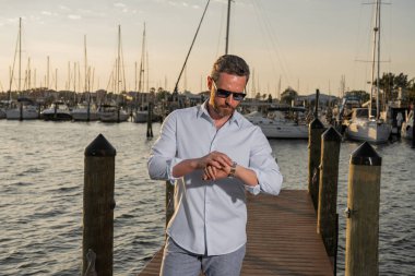 Yatın yanında zengin bir iş adamı var. Moda erkek modelinin açık havada portresi. Moda erkeği yatların yanında poz veriyor. Kentsel tarzda bir adam. Yat kulübündeki zengin adam. Deniz manzaralı ve lüks gemileri olan körfez adamı.