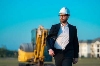 İnşaat alanında kask ve takım elbiseli bir inşaat yöneticisi. İnşaat müdürü ya da yönetici evin önünde şapka takıyor. Kazıcının yanındaki inşaat müdürü. Yenileme