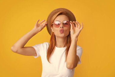 Young redhead woman in straw hat, surprised expression, isolated on yellow background. Summer lifestyle studio portrait. Redhead girl sends an air kiss