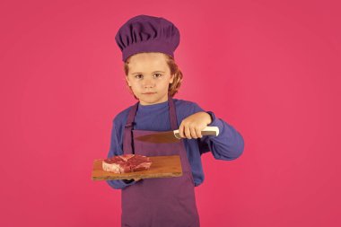 Child cook hold cutting board with meat beef steak and knife. Cooking, culinary and kids. Little boy in chefs hat and apron on studio isolated background