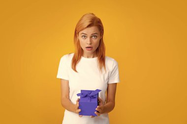 Surprised woman holding gift. Beautiful young woman in shirt hold present box with gift ribbon bow isolated on bright yellow background, studio portrait. Birthday concept