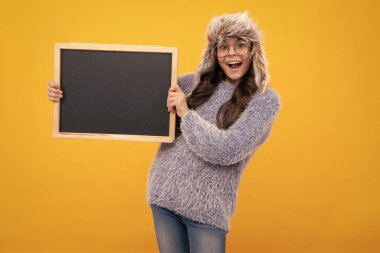 Teenage girl child in warm winter hat and sweater holding blackboard, isolated on a yellow background. Excited face, cheerful emotions of teenager girl