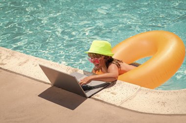 Havuzda rahatlayan bir çocuk, yaz suyunda dizüstü bilgisayar kullanıyor. Çocuk online çalışma ya da tropikal deniz plajında çalışıyor. Yaşam konsepti teknolojisi