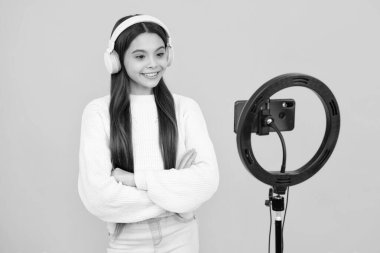 Music kids blog. Teenager child blogger with phone recording video on isolated yellow studio background. Influencer teen girl speaking in front of smartphone