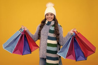 Sarı arka planda alışveriş torbaları olan, kışlık sıcak şapkalı, modaya uygun bir genç kız. Alışverişte çocuk alışverişi. Alışveriş çantalı çocuk.