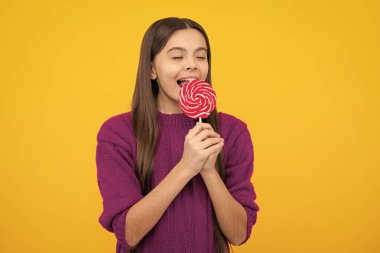 Funny child with lollipop over yellow isolated background. Sweet childhood life. Teen girl with yummy caramel lollipop, candy shop. Teenager with sweet sucker