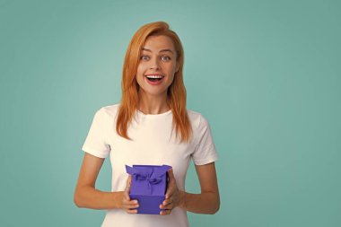 Elinde hediye tutan sürpriz bir kadın. Gülümseyen kızıl saçlı güzel bir kadın hediye kurdelesiyle hediye kutusunu tutuyor parlak mavi arka planda izole edilmiş, stüdyo portresi. Doğum günü kavramı