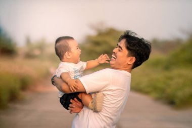 Babası, bebeği kaldırırken bebeğe sıcak bir şekilde gülümsedi. Baba, bebeği evin dışında tutuyor. Öğleden sonra doğal atmosferin arka planı.