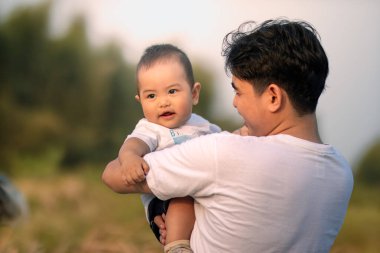 Bir erkek bebek babası tarafından kucaklanıyor ve baba bir gülümsemeyle bebeğe sıcak bir şekilde bakıyor. Baba, çocuğu açık alanda kameraya dönük bir şekilde tutuyor..