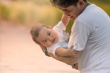 Baba, bebeği kucağına alıp kameraya gülümsüyor. Baba ve bebek beyaz tişört giyiyor. Öğleden sonra güneş ışığıyla doğal arka planda çekilmiş fotoğraf..