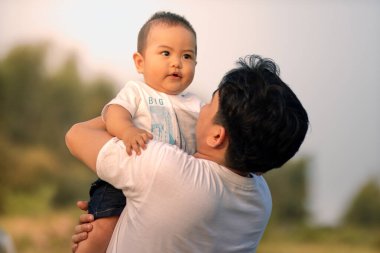 Bir erkek bebek gökyüzüne yükselirken kollarında tutuluyor. Baba, çocuğu açık alanda kameraya dönük bir şekilde tutuyor..