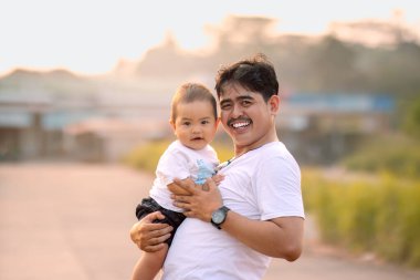 Baba, bebeği sıcak bir şekilde tutuyor ve çocuğun elini tutarak kameraya gülümsüyor. Erkek bebek diğer yönde sevimli bir gülümseme takındı..